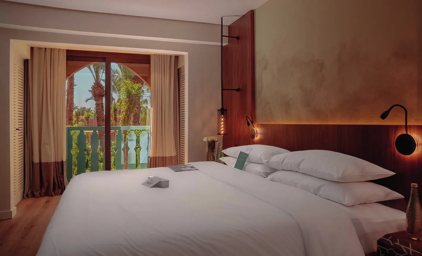 Deluxe room: large white bed and an arched French window opening onto the balcony and palms